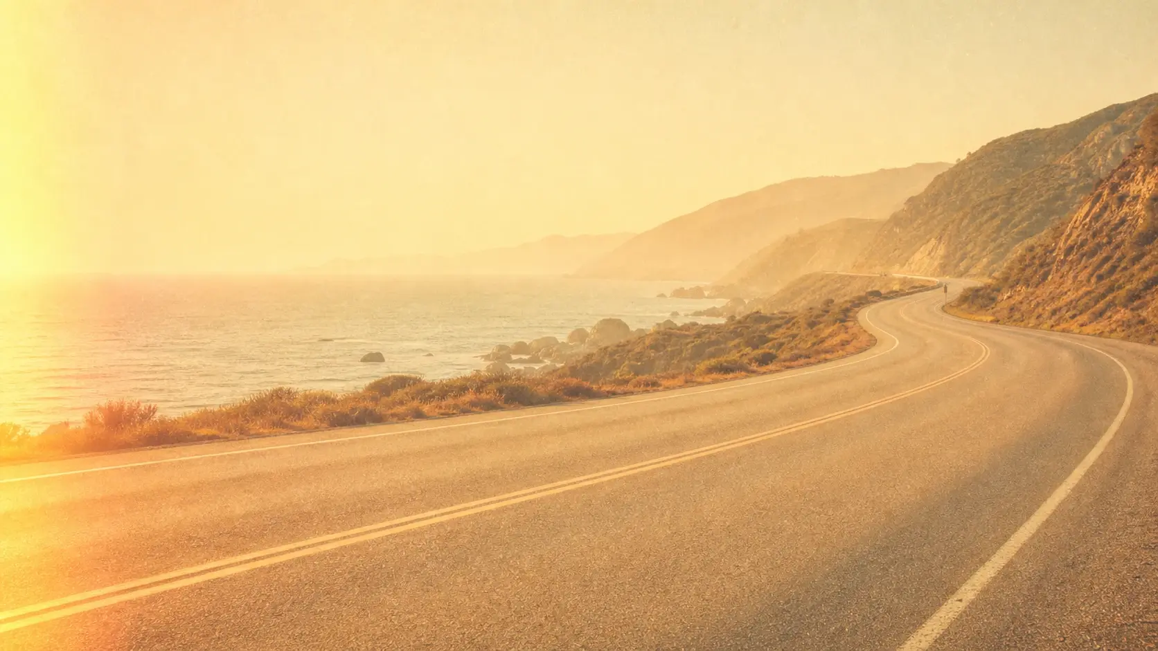 A wide sunlit coastal road rendered as warm faded film with soft grain and a gentle light leak